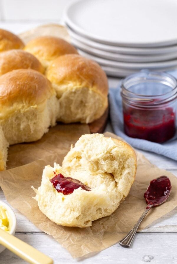A vertical image showing the sourdough discard rolls in the background of the photo, along with a stack of white plates. In the foreground, there is a sourdough roll that has been broken open and spread with some of the strawberry jam from the jar sitting beside it.