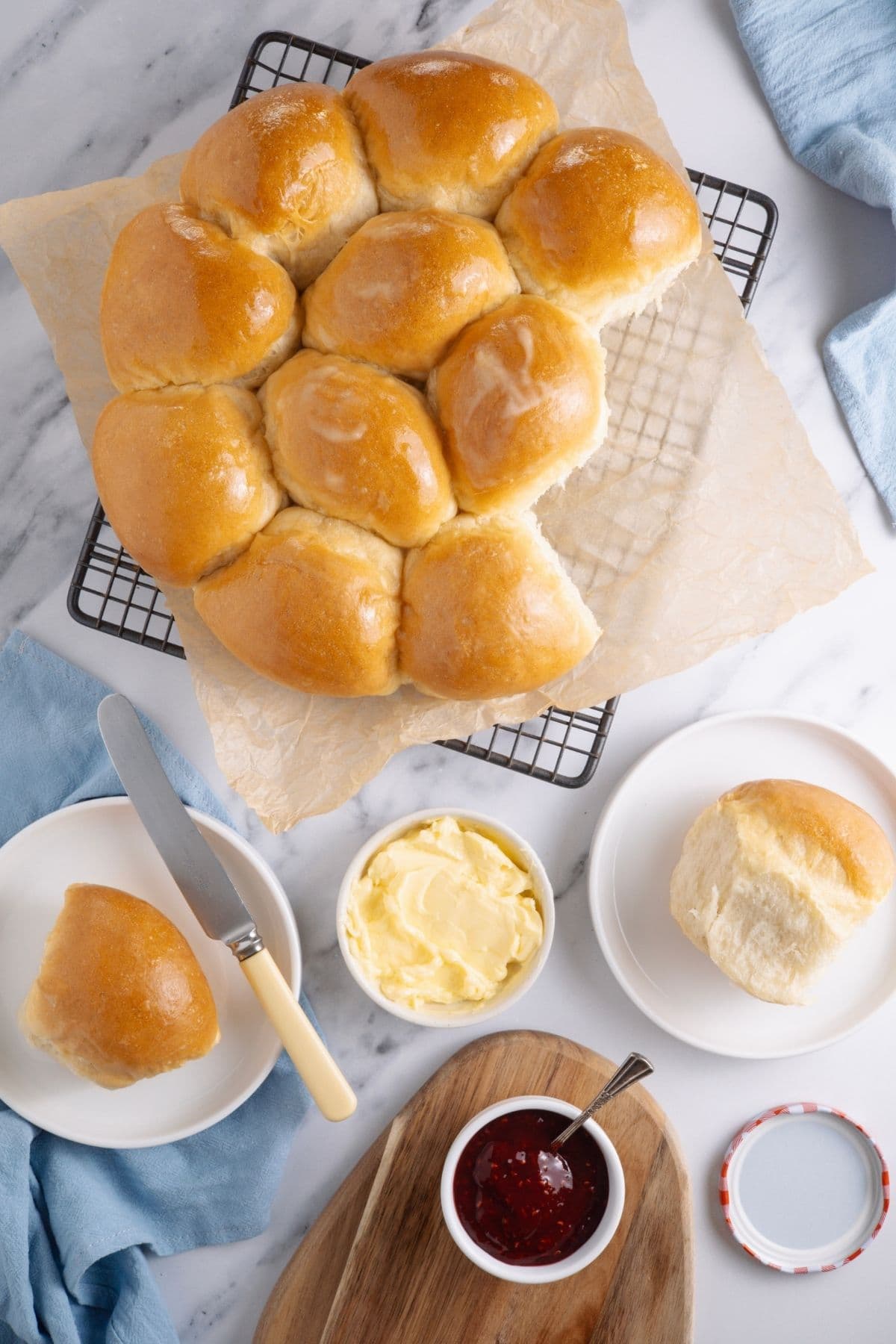 Flat lay image of sourdough discard rolls that have just come out of the oven. They are cooling on a wire rack and there is a small pot of strawberry jam and butter, ready to be spread onto the warm rolls. There are some pale blue linen napkins placed around the edges of the photo too.