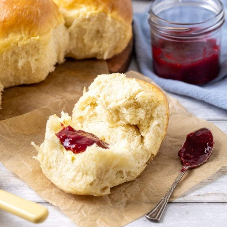 An image showing the sourdough discard rolls in the background of the photo, along with a stack of white plates. In the foreground, there is a sourdough roll that has been broken open and spread with some of the strawberry jam from the jar sitting beside it.