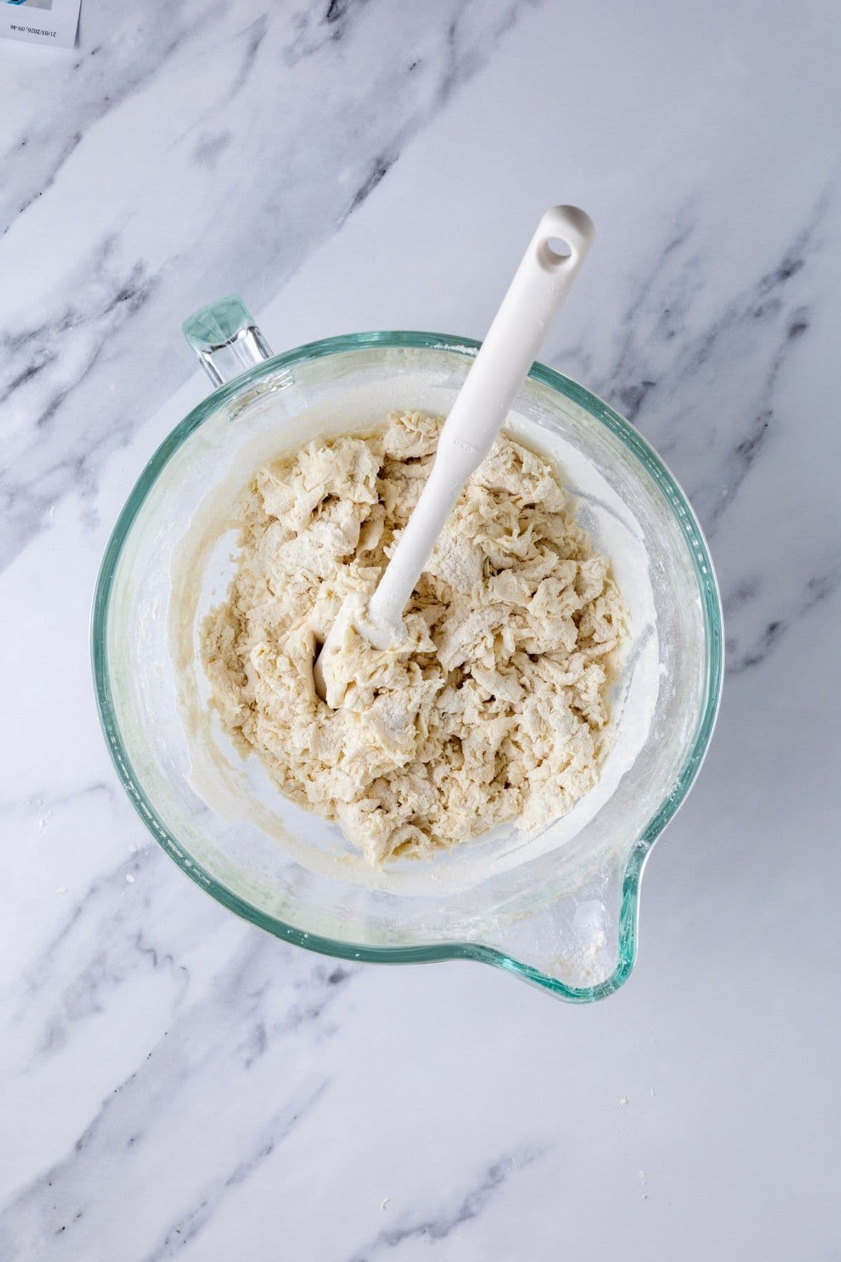 A glass bowl from a stand mixer containing the dry, rough dough from the initial mix of ingredients for making sourdough discard rolls.