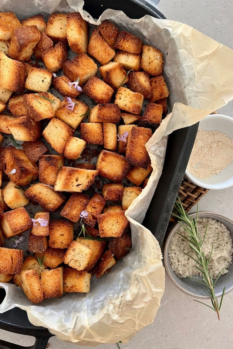 Crunchy homemade sourdough croutons in a cast iron dish lined with parchment paper. There is a bowl of french sea salt and sprig of rosemary to the right.