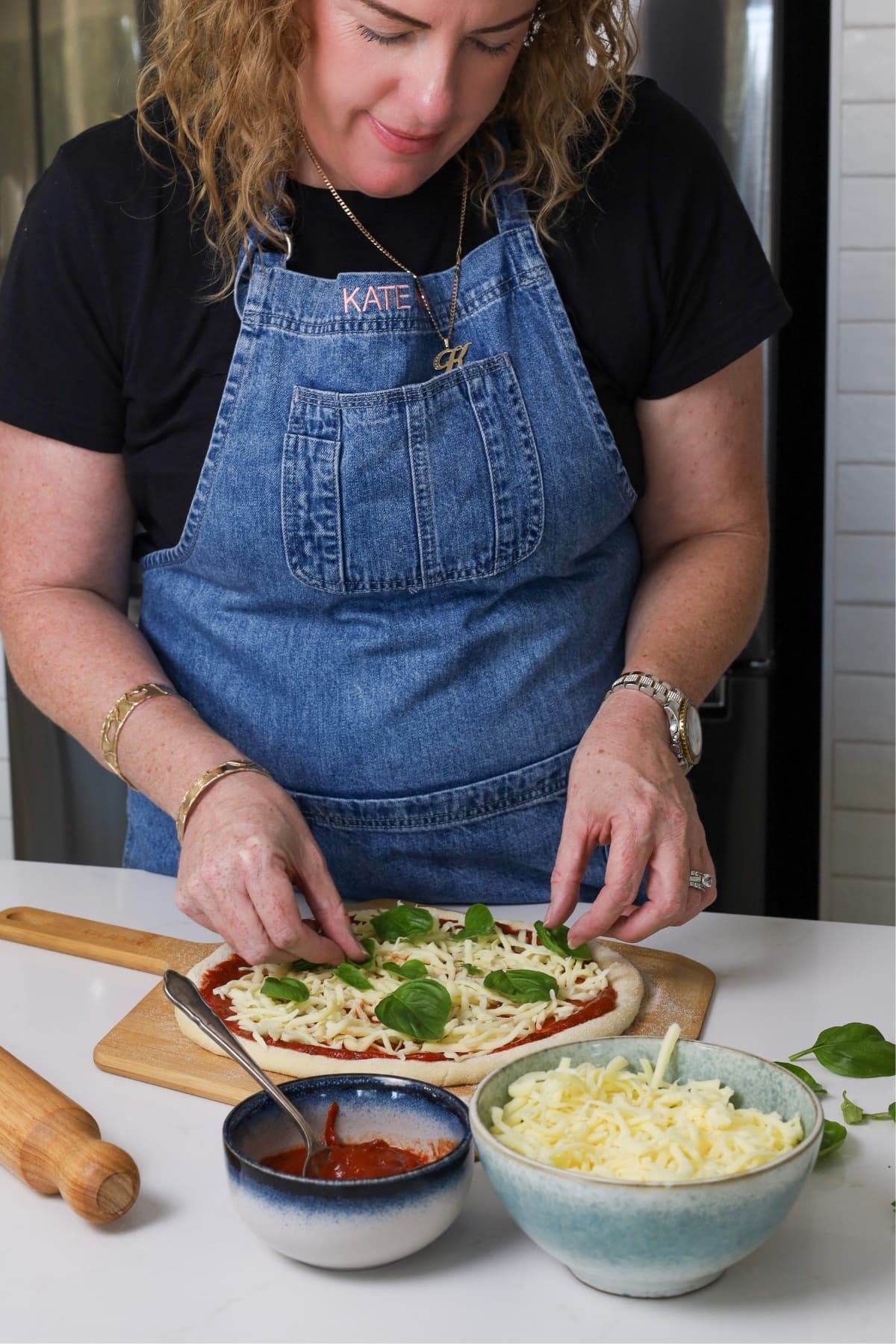 Kate from The Pantry Mama adding mozzarella and basil to a sourdough discard pizza while its sitting on a wooden pizza peel. Kate is wearing a black t-shirt and denim apron.