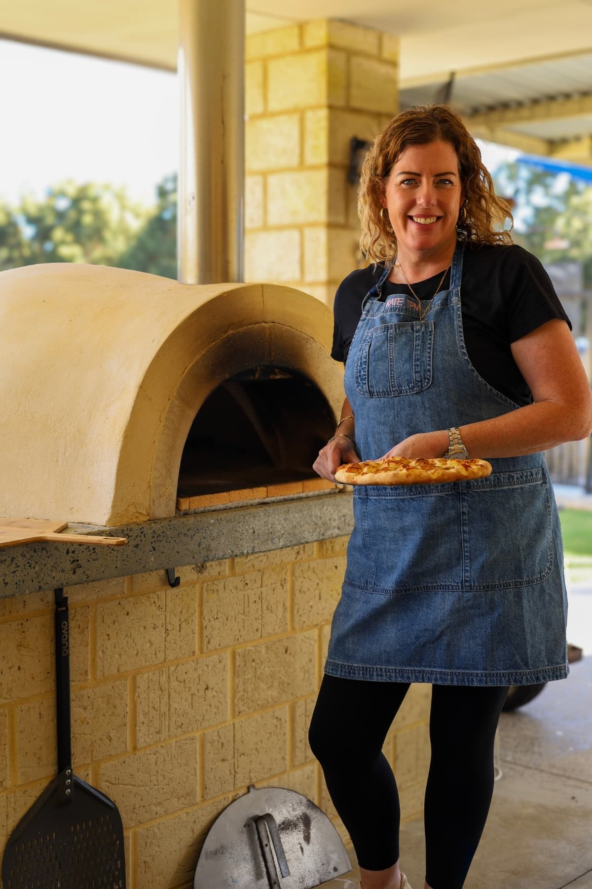 Kate from The Pantry Mama standing in front of a wood fired pizza oven holding a pizza peel. There is a freshly baked sourdough discard pizza on the peel. Kate is wearing black clothes with a denim apron over the top.