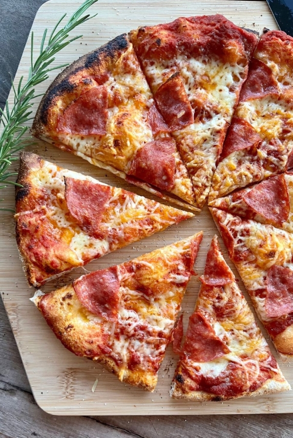 Sourdough pizza topped with pepperoni and cheese and sitting on a wooden pizza peel. There is a sprig of roseary on the left side of the pizza.