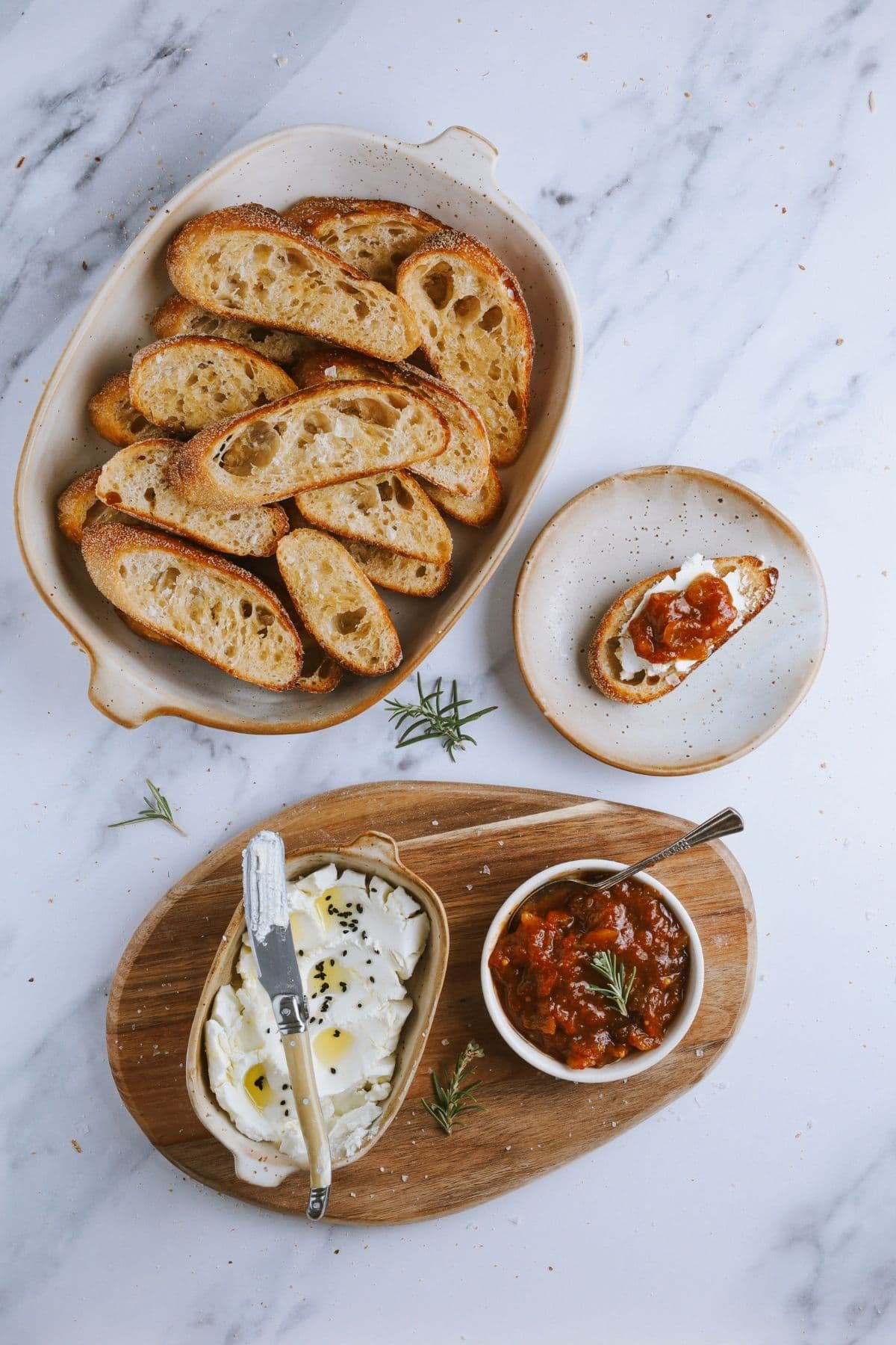 A flat lay showing a plate of perfectly toasted crostini next to a wooden board holding a dish of goats cheese and a dish of tomato chutney. There is a third plate in the scene showing a sourdough crostini topped with goats cheese and relish.