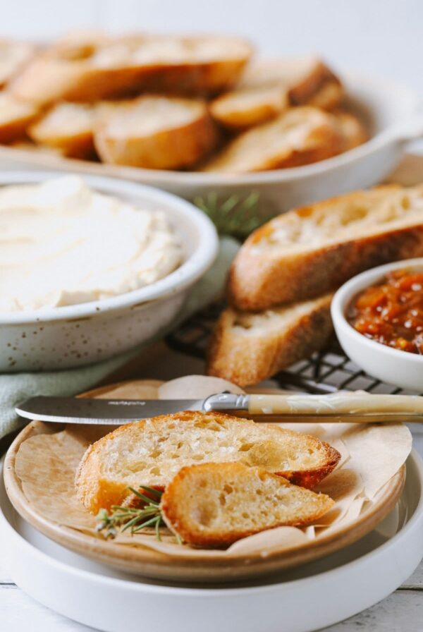 A beautiful display of sourdough crostini sitting on small white plate. You can see some whipped honey cream cheese and tomato chutney in the background.