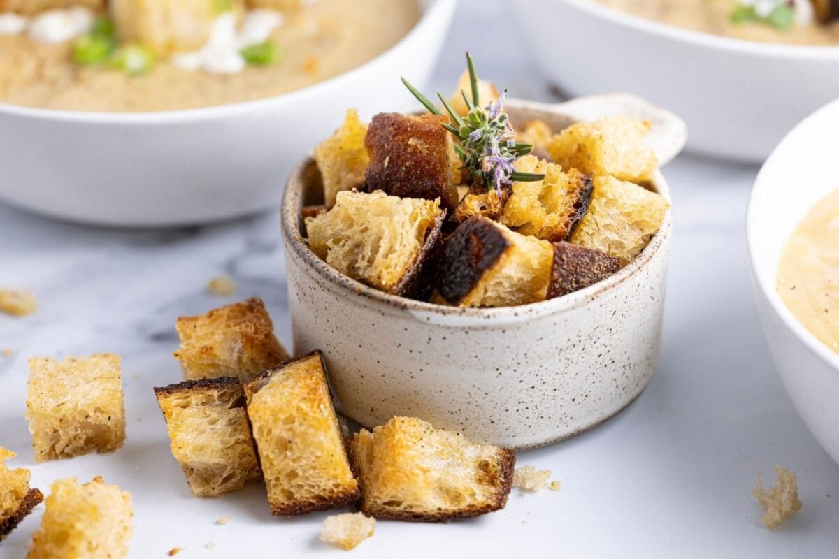 A horizontal image of a small speckled dish filled with golden brown sourdough croutons. The croutons are spilling out onto the counter. In the background you can see bowls of vegetable soup.