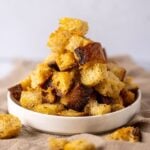Recipe feature image for Sourdough Croutons showing a small white dish filled with golden brown sourdough croutons spilling out onto the counter.
