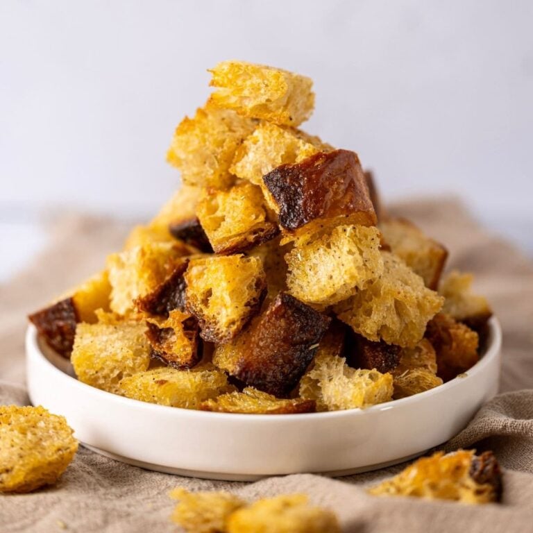 Recipe feature image for Sourdough Croutons showing a small white dish filled with golden brown sourdough croutons spilling out onto the counter.