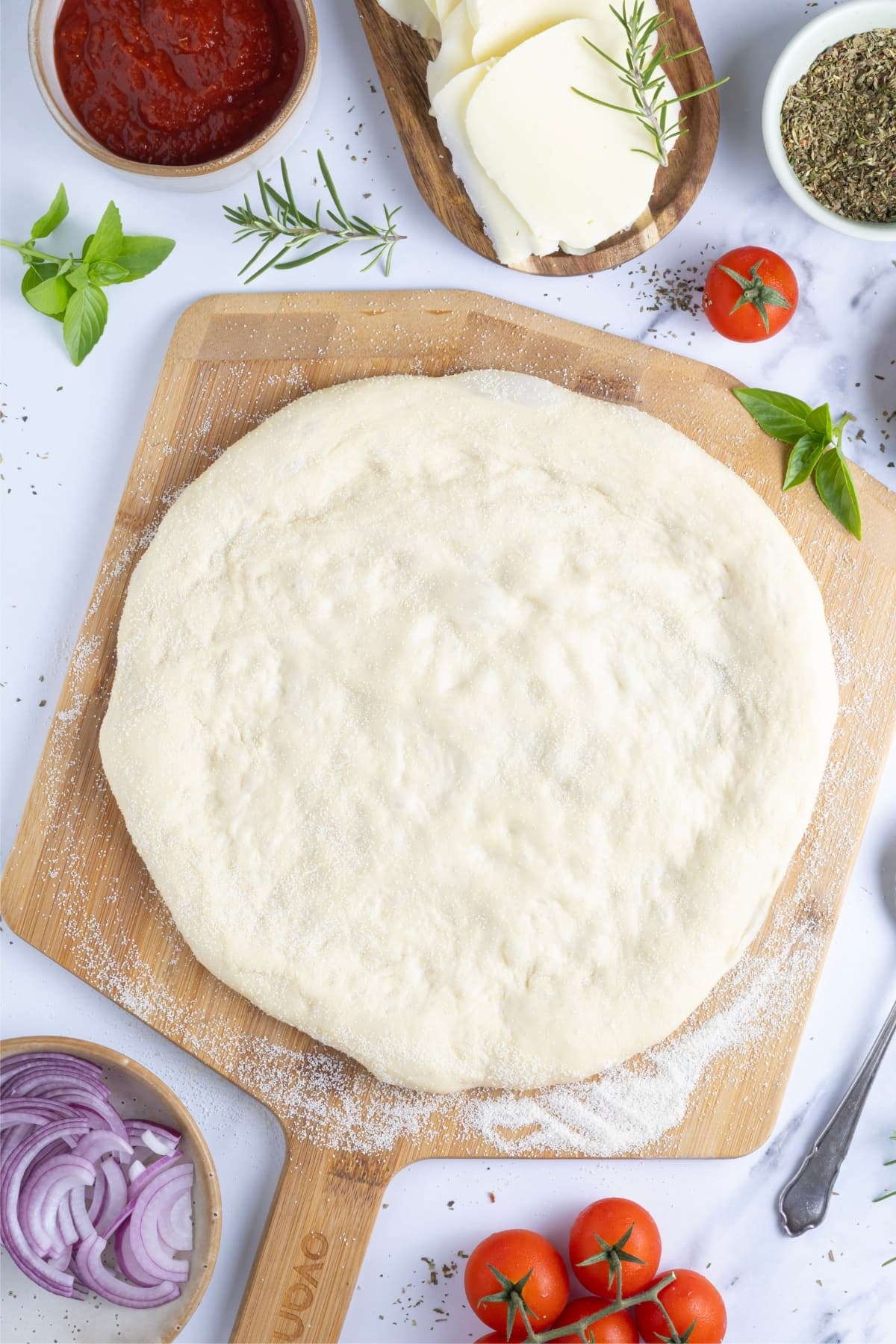 A round sourdough discard pizza dough crust sitting on a wooden pizza peel. Surrounding the pizza peel are small bowls and trays containing pizza toppings like fresh mozzarella, red onion and cherry tomatoes.