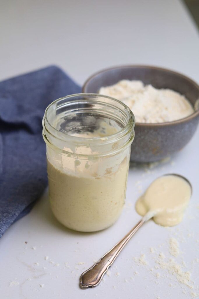 A small jar of sourdough starter sitting next to a spoon that has sourdough starter dripping from it. There is also a bowl of flour and blue dish towel in the background of the photo.