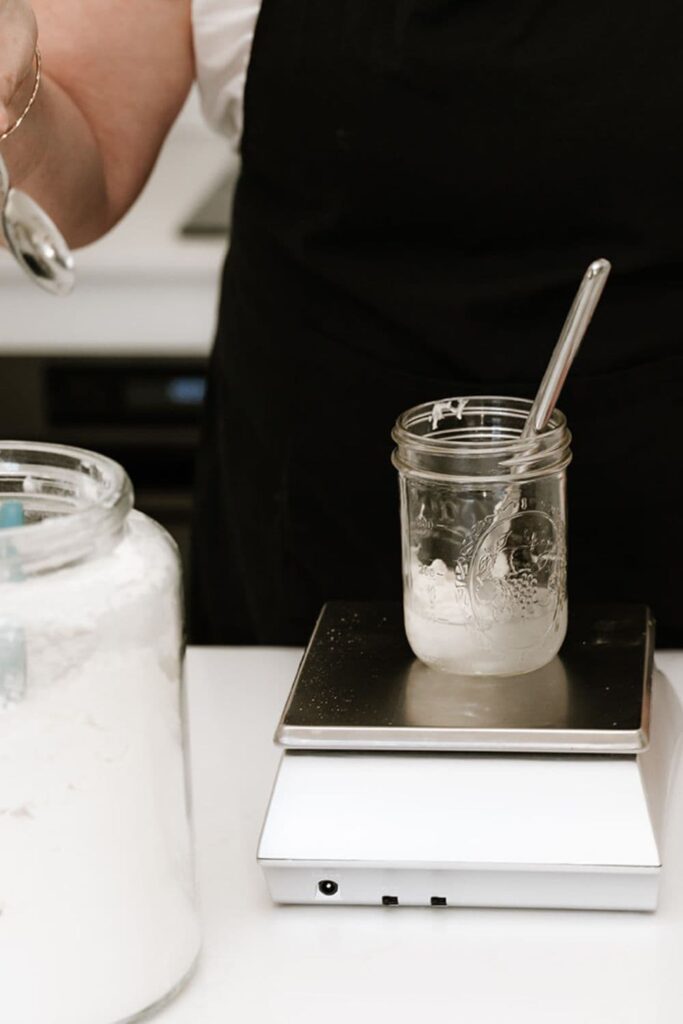 A jar of sourdough starter sitting on top of a digital scale. There is a knife in the jar that will be used to stir the starter and a jar of flour in the photo too.