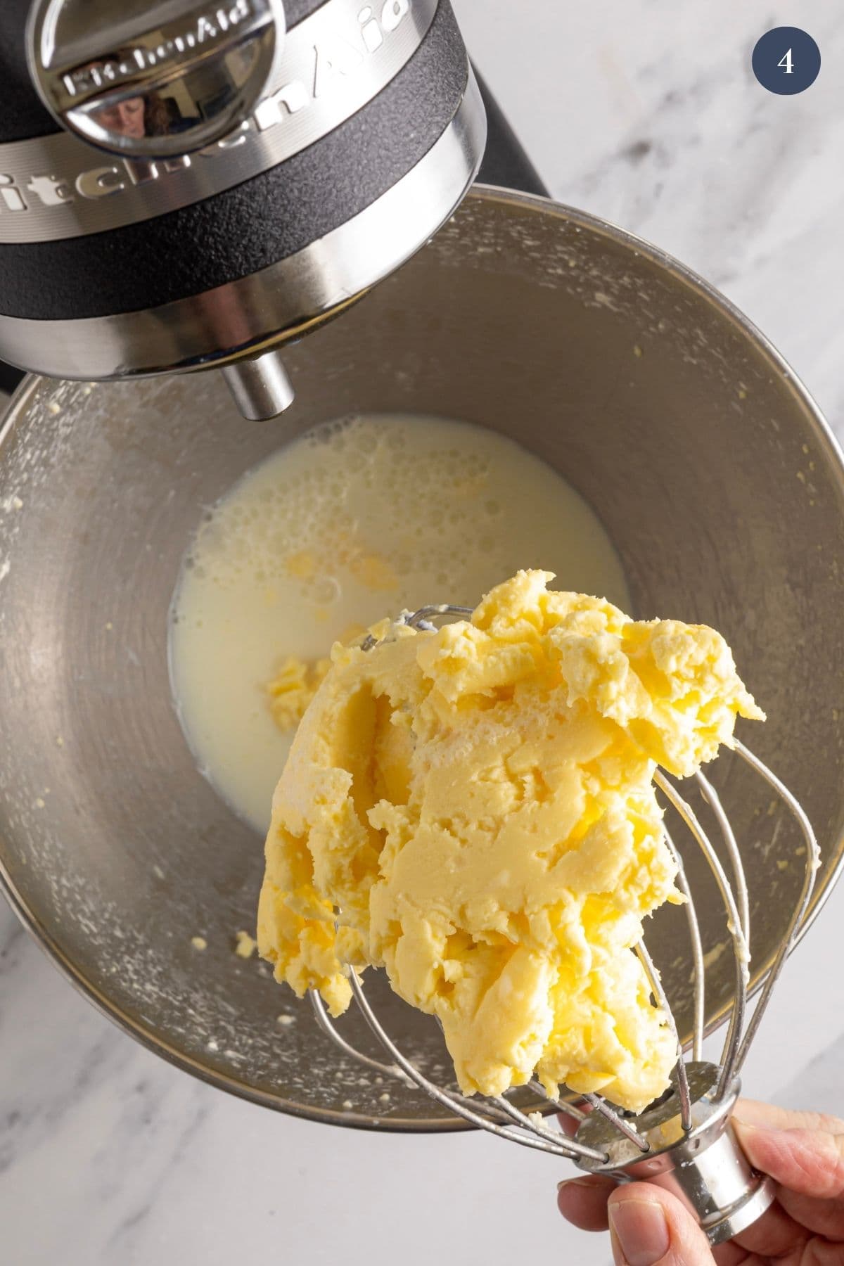 A photo taken from above showing the cream that has separated into buttermilk and butter. The butter has accumulated in the wire whisk attachment of the stand mixer and is being held over the edge of the bowl to demonstrate the two elements.