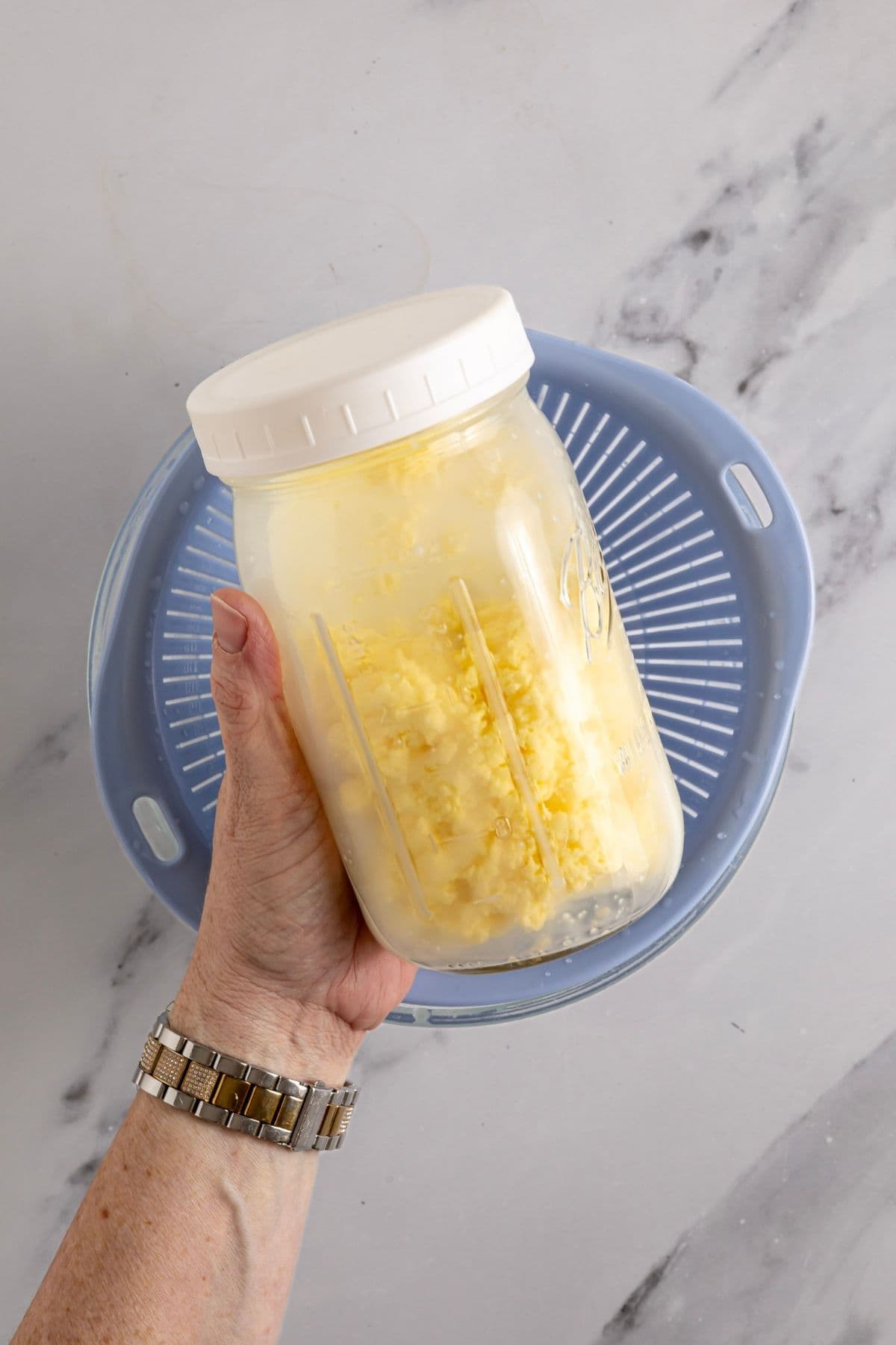 A hand holding a 32oz jar that contains butter that has just been shaken from a pint of cream. There is a blue strainer shown behind the jar, ready to strain out the buttermilk.
