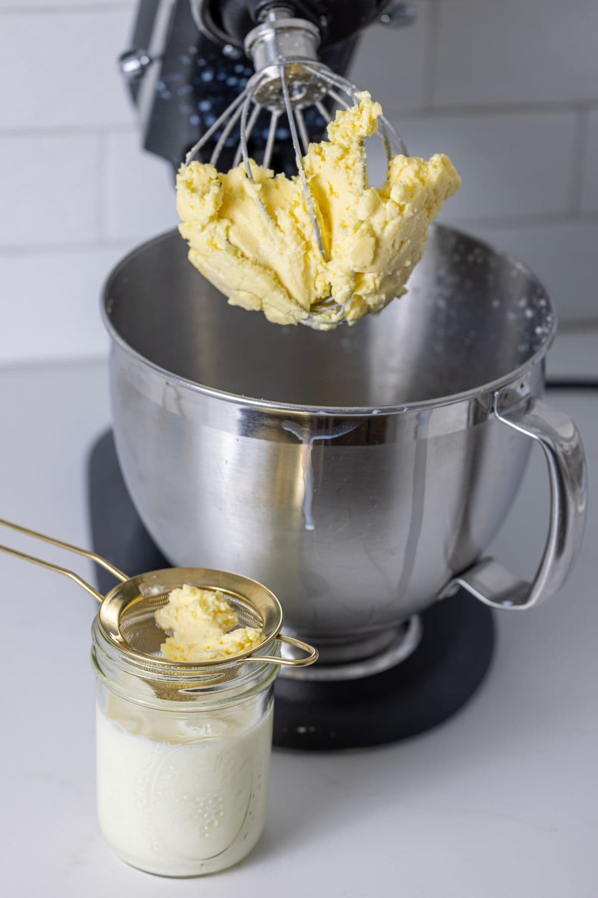 A vertical image showing a KitchenAid with the butter held in the whisk attachment. There is a small jar of buttermilk next to the mixer with a small strainer on top that has caught some of the butter.