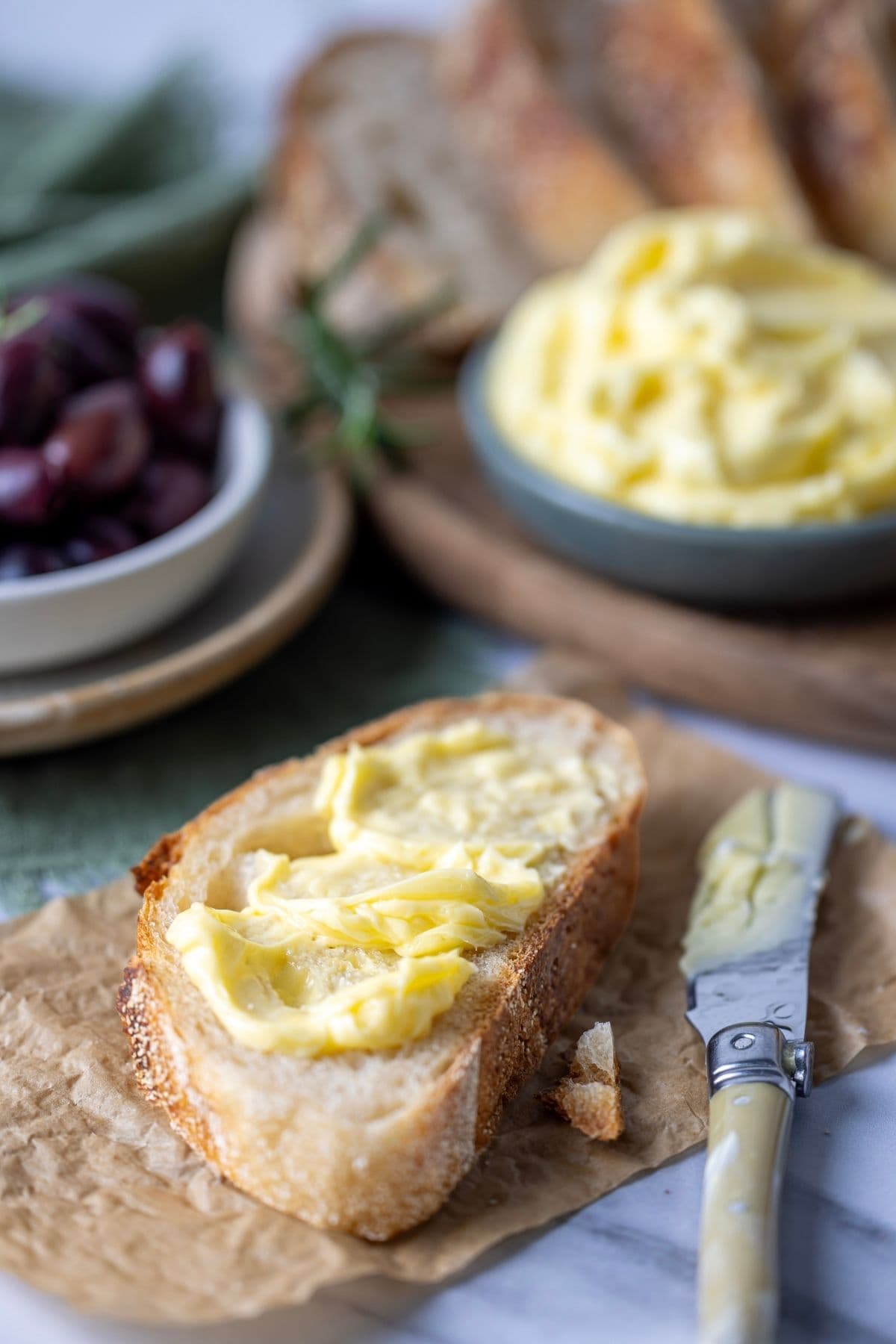 Cultured butter spread onto a slice of sourdough bread. There is a dish of cultured butter and a dish of Kalamata olives in the background and a butter knife laying next to the slice of bread.