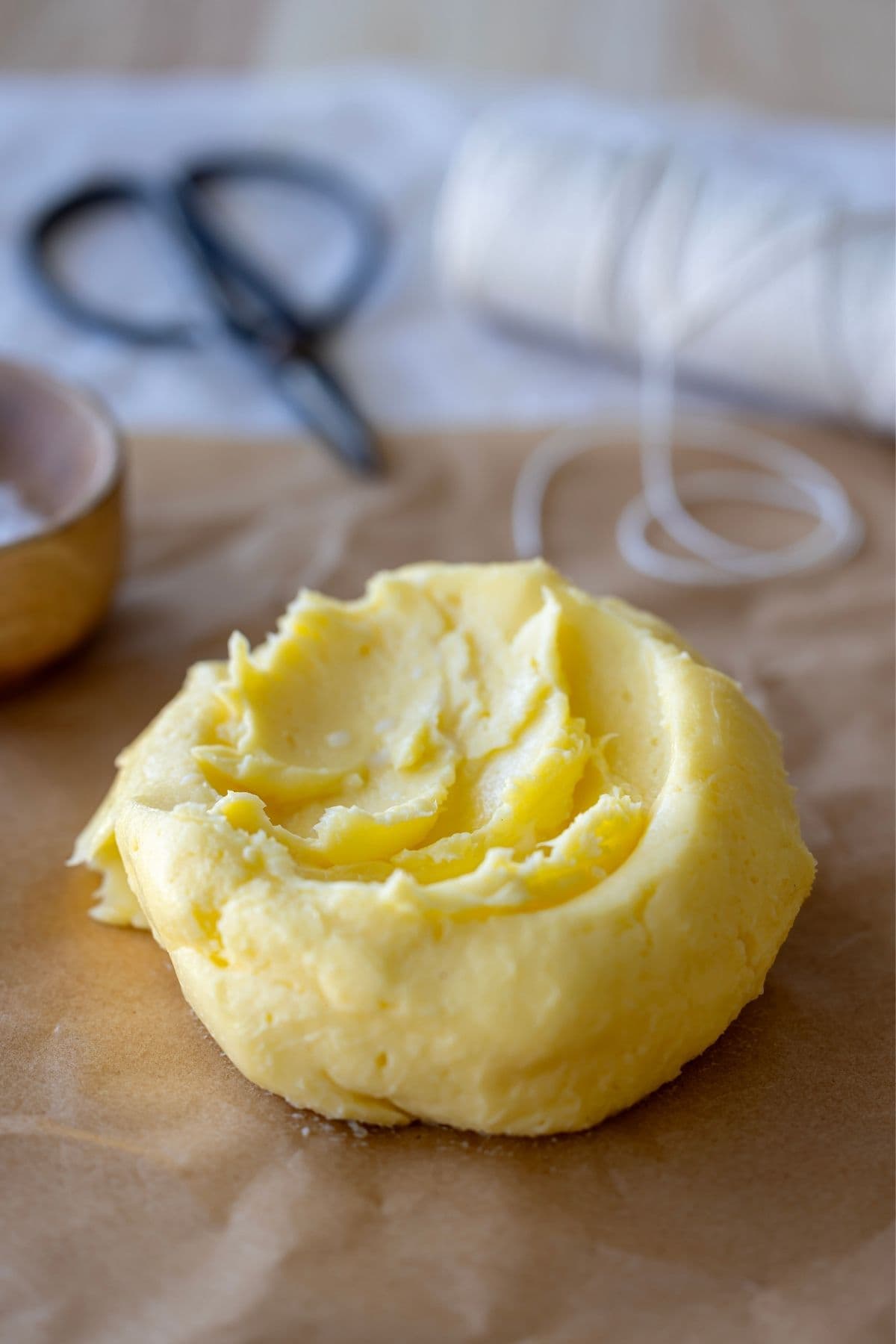 A batch of homemade butter shaped into a round. it is sitting on a piece of parchment paper ready to be wrapped up. There is string and scissors in the background of the photo ready to tie up the package.