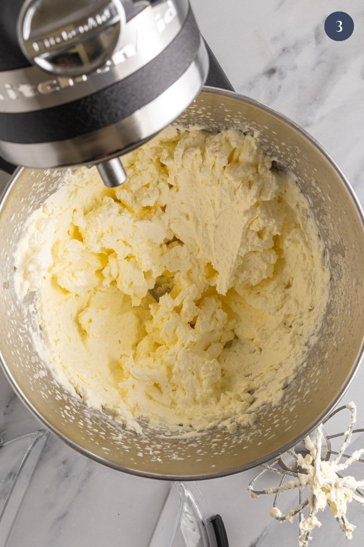 The bowl of a stand mixer showing the grainy, curdled looking whipped cream that is on the way to becoming butter.