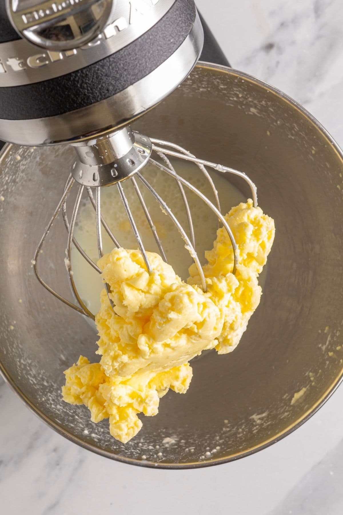 A photo taken from above showing the cream that has separated into buttermilk and butter. The butter has accumulated in the wire whisk attachment of the stand mixer and you can see the cloudy buttermilk in the bottom of the bowl.