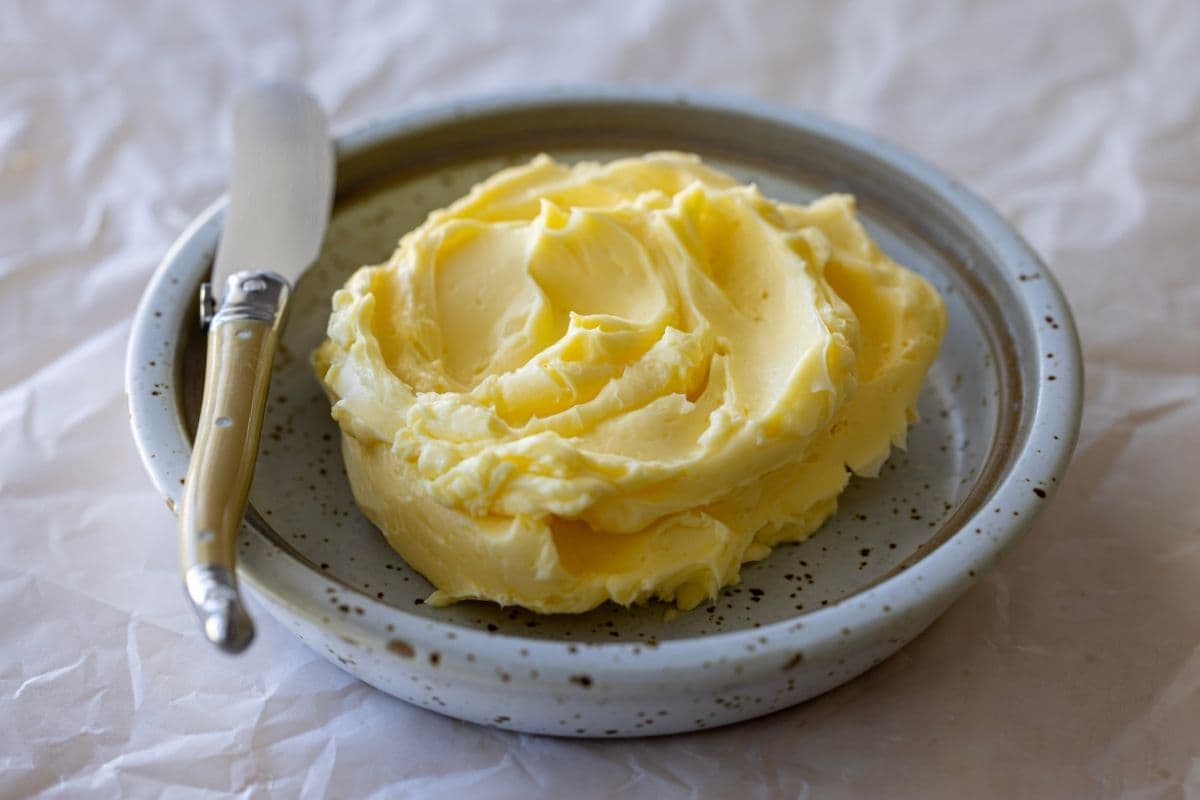 A swirl of homemade butter sitting in a stoneware butter dish. There is a butter spreading knife sitting on the edge of the dish.