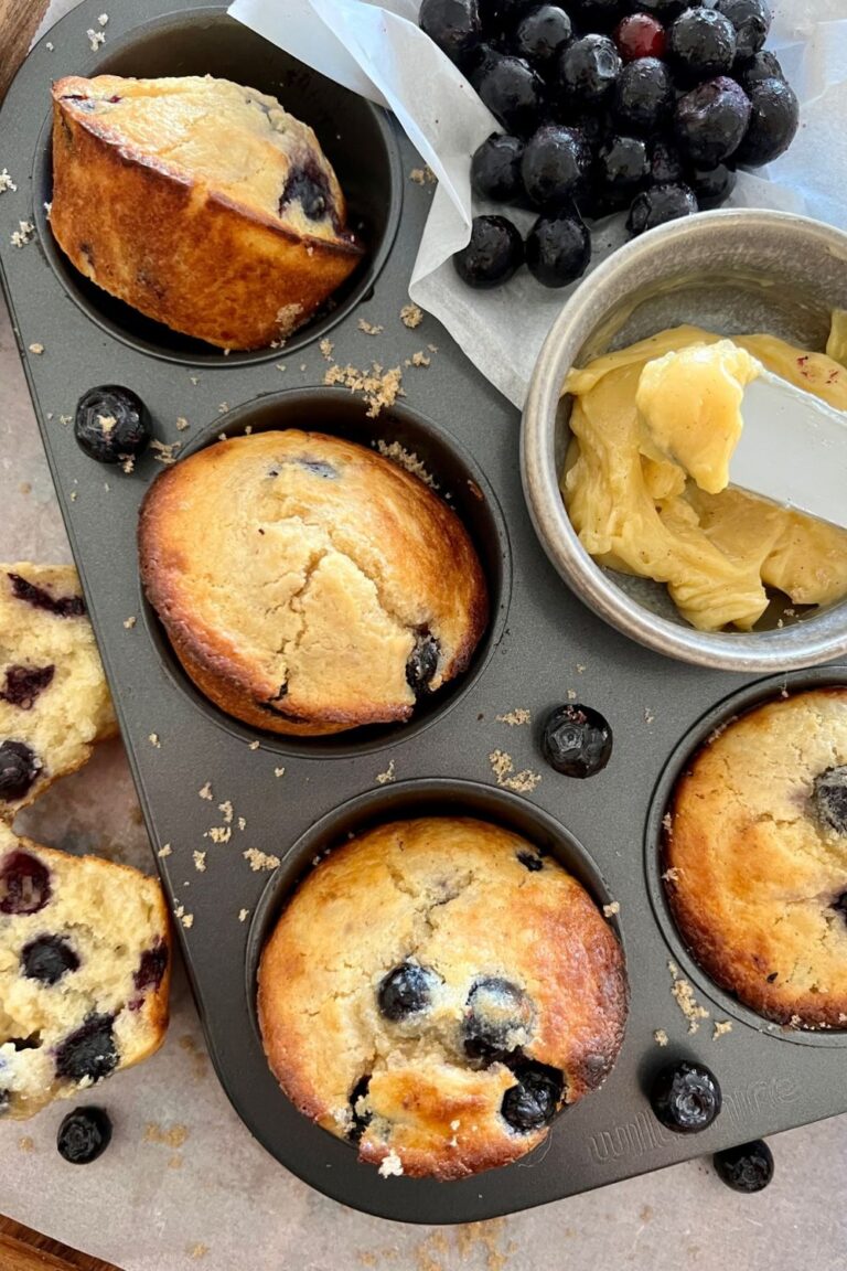 A 6 hole muffin tray containing sourdough blueberry muffins that have just been baked. There is also a pile of fresh blueberries in the top right of the photo.