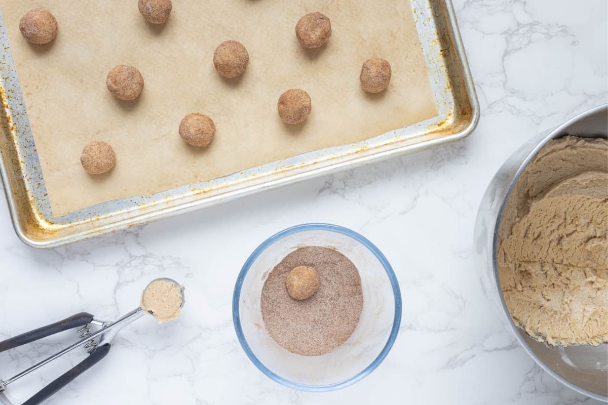 A metal tray lined with parchment paper holding balls of sourdough snickerdoodle dough. There is also a cookie scoop with some dough in it and a small bowl of cinnamon sugar mixture that has a ball of dough being rolled in it.