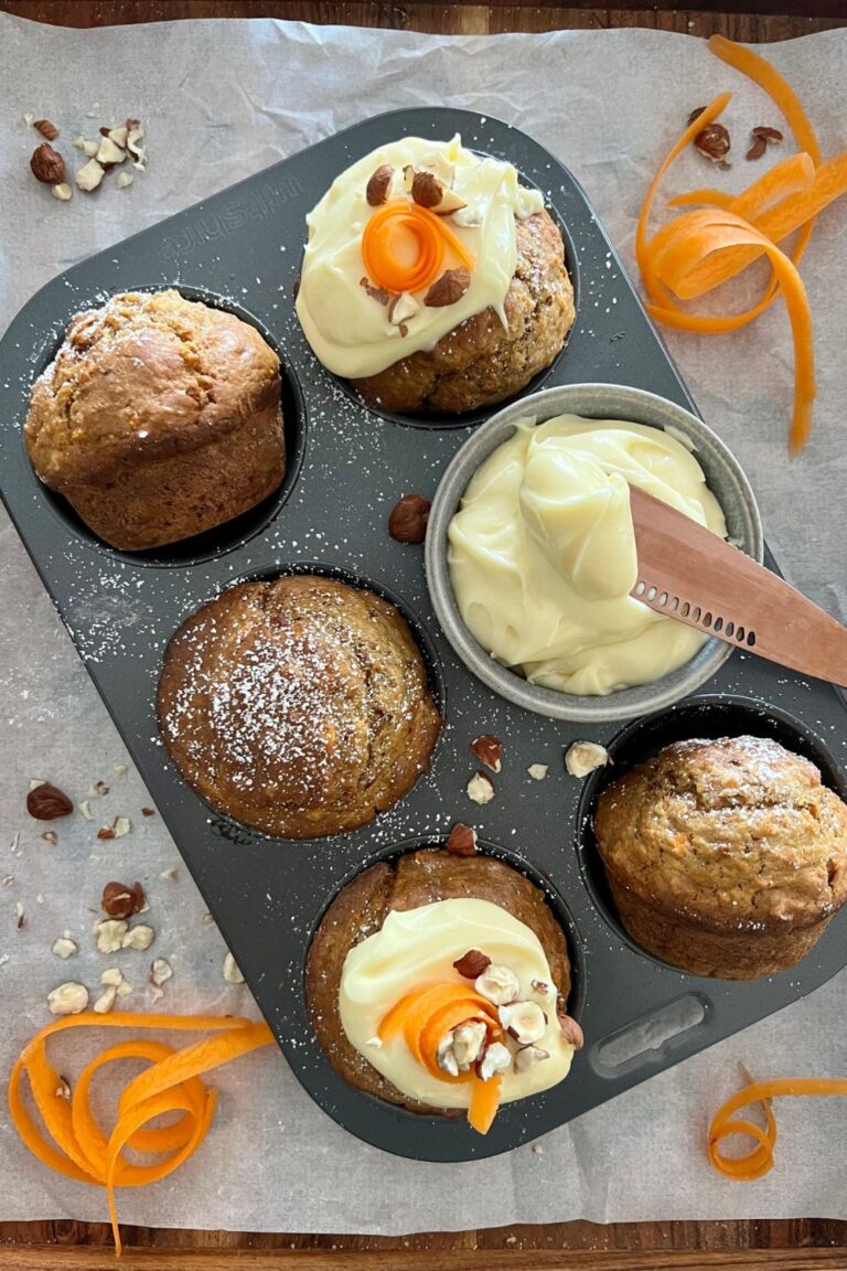 Sourdough carrot cake muffins in a six cup muffin tray. Two of them are topped with cream cheese frosting and carrot garnish.