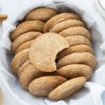 A batch of sourdough snickerdoodle cookies in a round container lined with white linen. There is a bite taken out of the top snickerdoodle cookie.