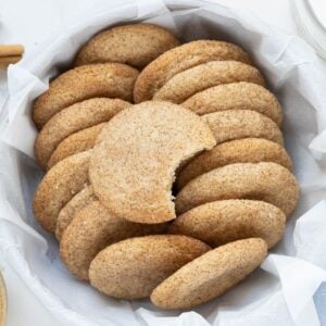 A batch of sourdough snickerdoodle cookies in a round container lined with white linen. There is a bite taken out of the top snickerdoodle cookie.