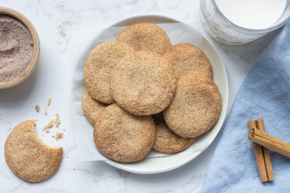 A batch of sourdough snickerdoodle cookies arranged on a white plate. There is a glass of milk next to the plate and a snickerdoodle with a bite taken out of it on the left.