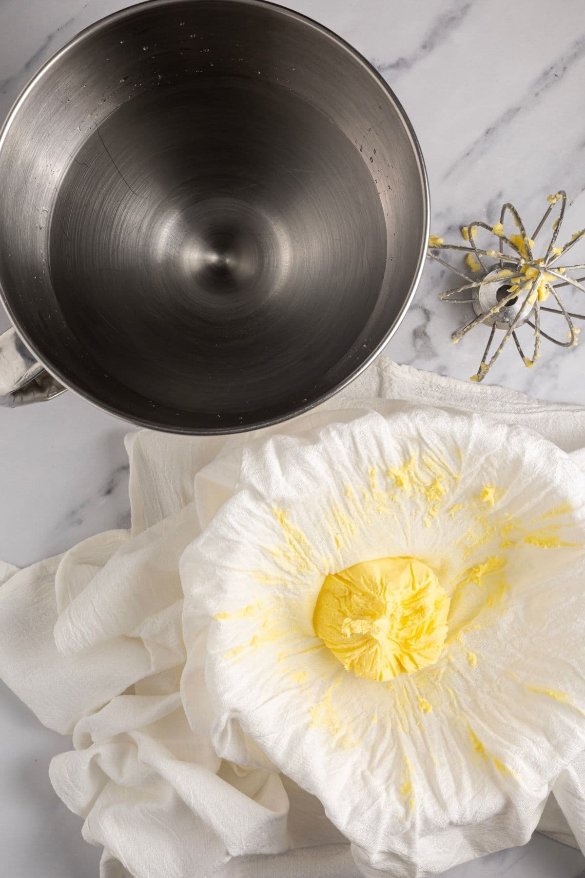A photo showing the butter being squeezed in some fine cheesecloth next to a stand mixer bowl filled with clean water ready to wash the butter again.