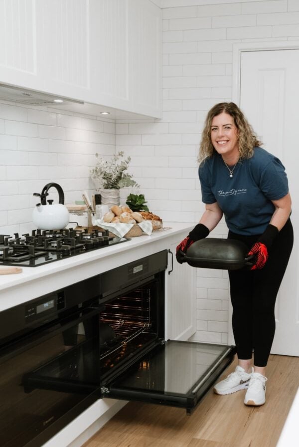 Kate, the founder of The Pantry Mama, putting a cast-iron bread pan into a large electric oven. The oven door is open so you can see inside the oven. Kate is wearing a blue t-shirt. There is a white kettle on the gas cook top on the top of the oven.