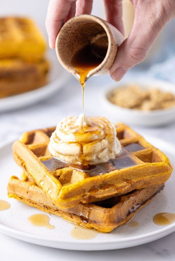 2 sourdough pumpkin waffles stacked on top of each other and topped with whipped ricotta. There is a hand holding a small jug and pouring maple syrup over the top.