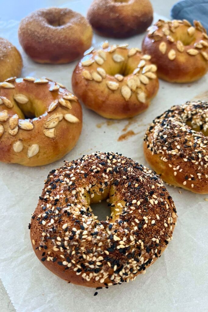 A selection of sourdough pumpkin bagels with different toppings. The ones at the front are covered in everything bagel seasoning, the middle ones are topped with pumpkin seeds and the ones at the back are dusted in cinnamon sugar.