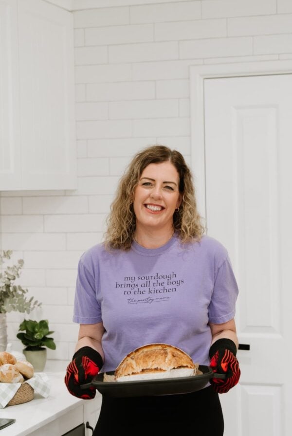A photo of Kate from The Pantry Mama wearing a purple t-shirt and holding a baked loaf of sourdough in a cast-iron bread pan demonstrating how to make great sourdough bread in heat and humidity.