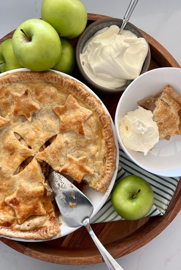Sourdough apple pie displayed on a round wooden tray. A slice has been cut from the apple pie and there is a silver pie slice sitting in the dish. The dish is surrounded by 5 green granny smith apples, a bowl of whipped cream and a dish with a serve of apple pie and whipped cream.