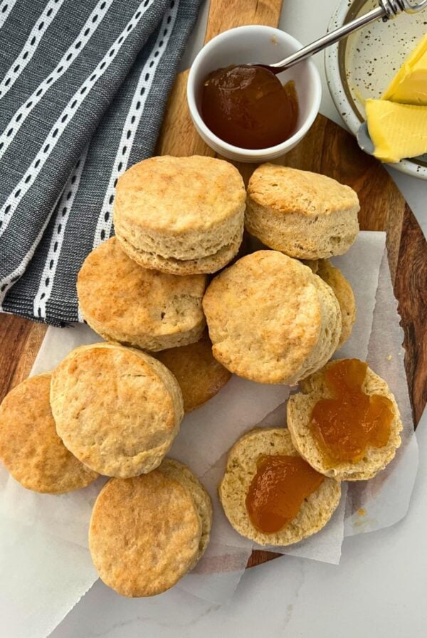 A photo of sourdough biscuits piled high on a wooden board and spread with apricot jam.
