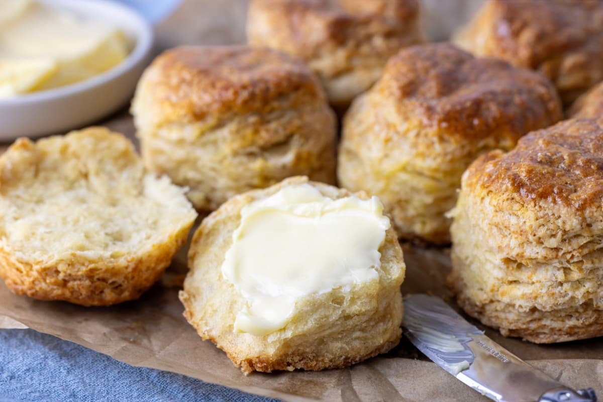 A batch of sourdough biscuits displayed on a sheet of parchment paper. One of the biscuits at the front has been split open and spread with butter.