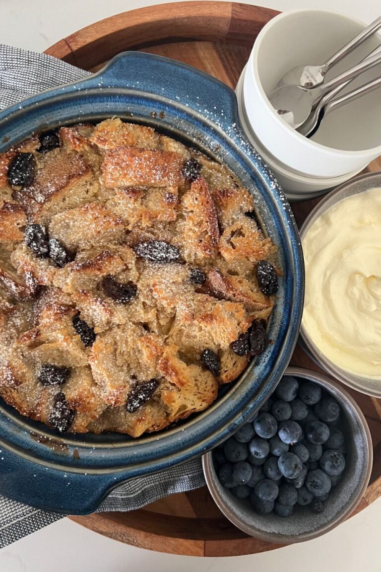 Sourdough bread pudding with raisins baked in a blue casserole dish. It is on a wooden tray with a dish of whipped cream and a bowl of blueberries.