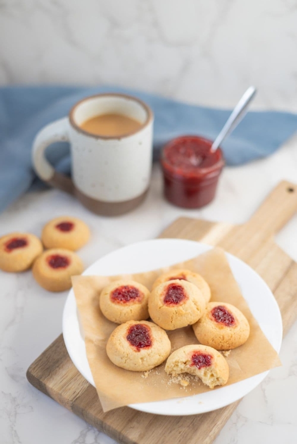 A white plate filled with sourdough discard thumbprint cookies. There is a cup of coffee and jar of strawberry jam in the background of the photo.