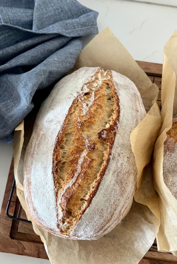 Loaf of rustic sourdough bread sitting in a piece of parchment paper on a wooden board. There is a blue dish towel on the left and you can see the edge of a second loaf of sourdough on the right.