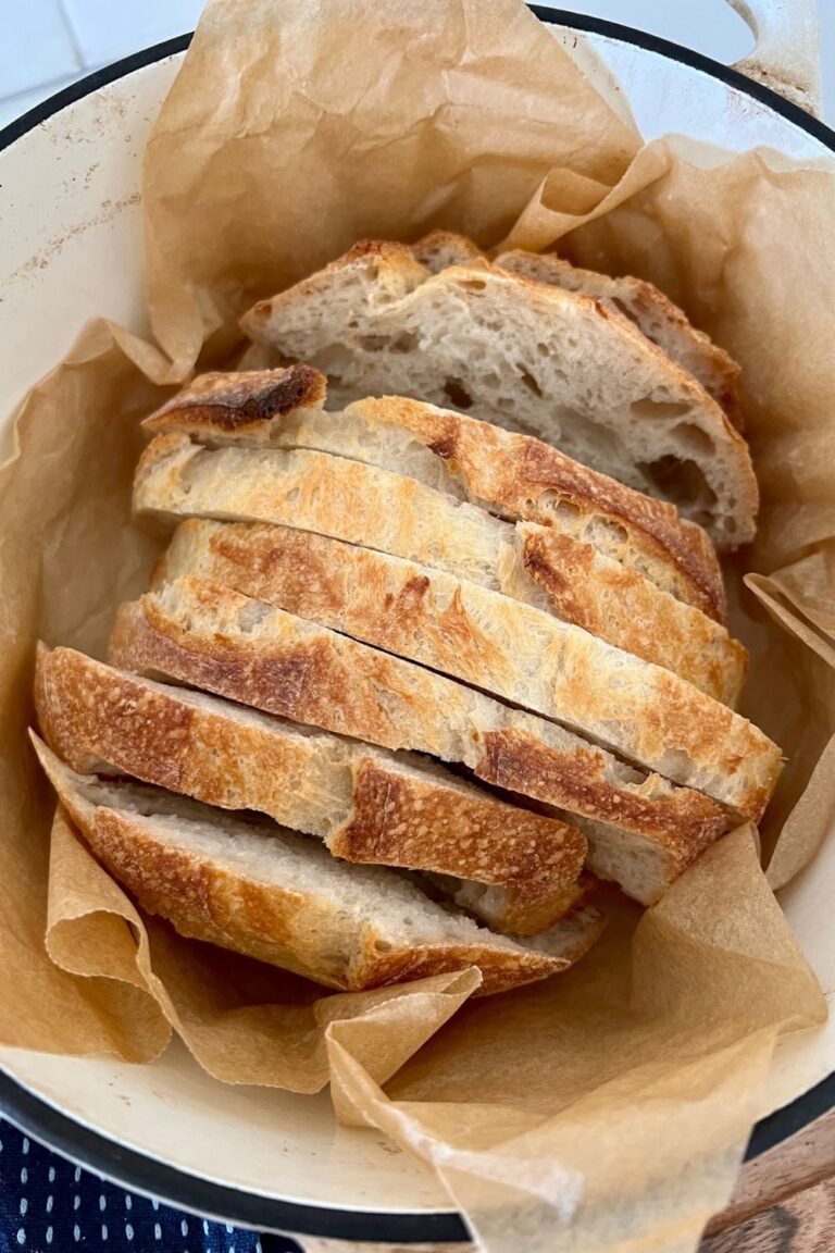 Small loaf of sourdough bread that has been sliced sitting in a cream enamel Dutch Oven surrounded by parchment paper.