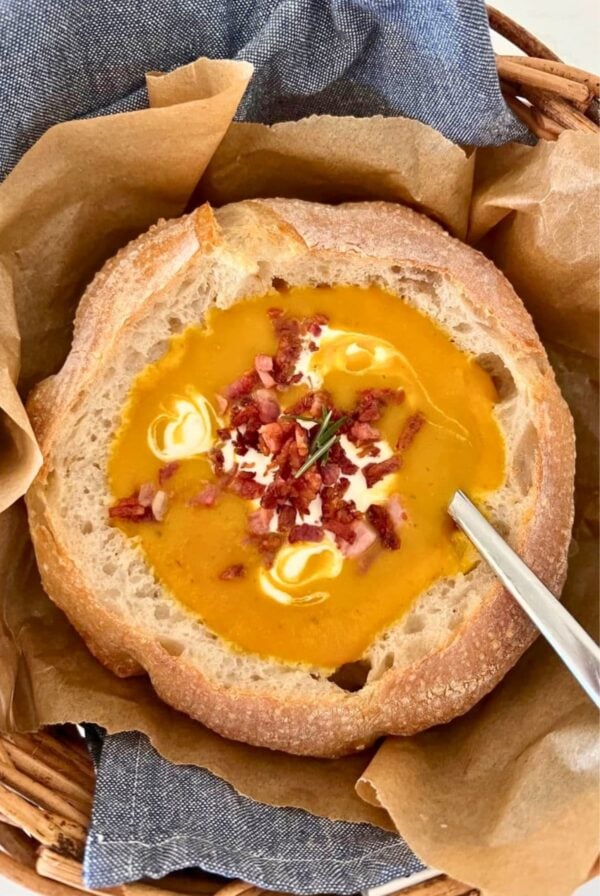 A sourdough bread bowl filled with bright orange pumpkin soup topped with sour cream, bacon pieces and parsley.