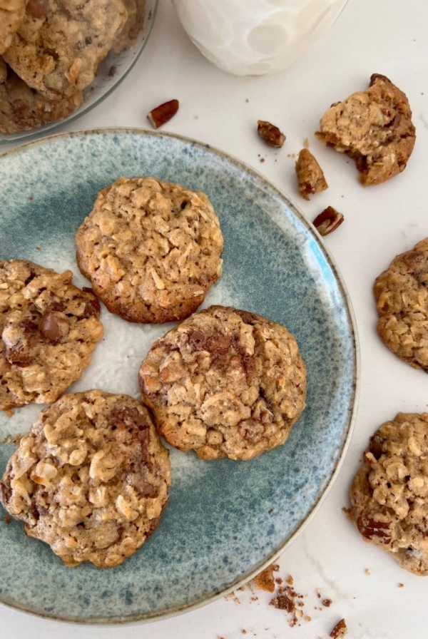 4 sourdough discard cowboy cookies on a blue stoneware plate. There is a glass of milk in the background and some cookie crumbs around the edges of the plate.