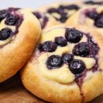 A selection of sourdough brioche blueberry cheesecake rolls sitting on a wooden board. The rolls are filled with vanilla cheesecake filling and topped with blueberries that have started to ooze out when cooked.