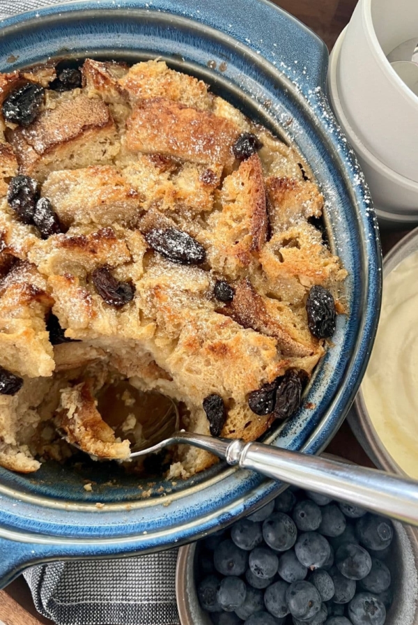 A blue casserole dish filled with sourdough bread pudding that is being scooped out with a silver spoon.