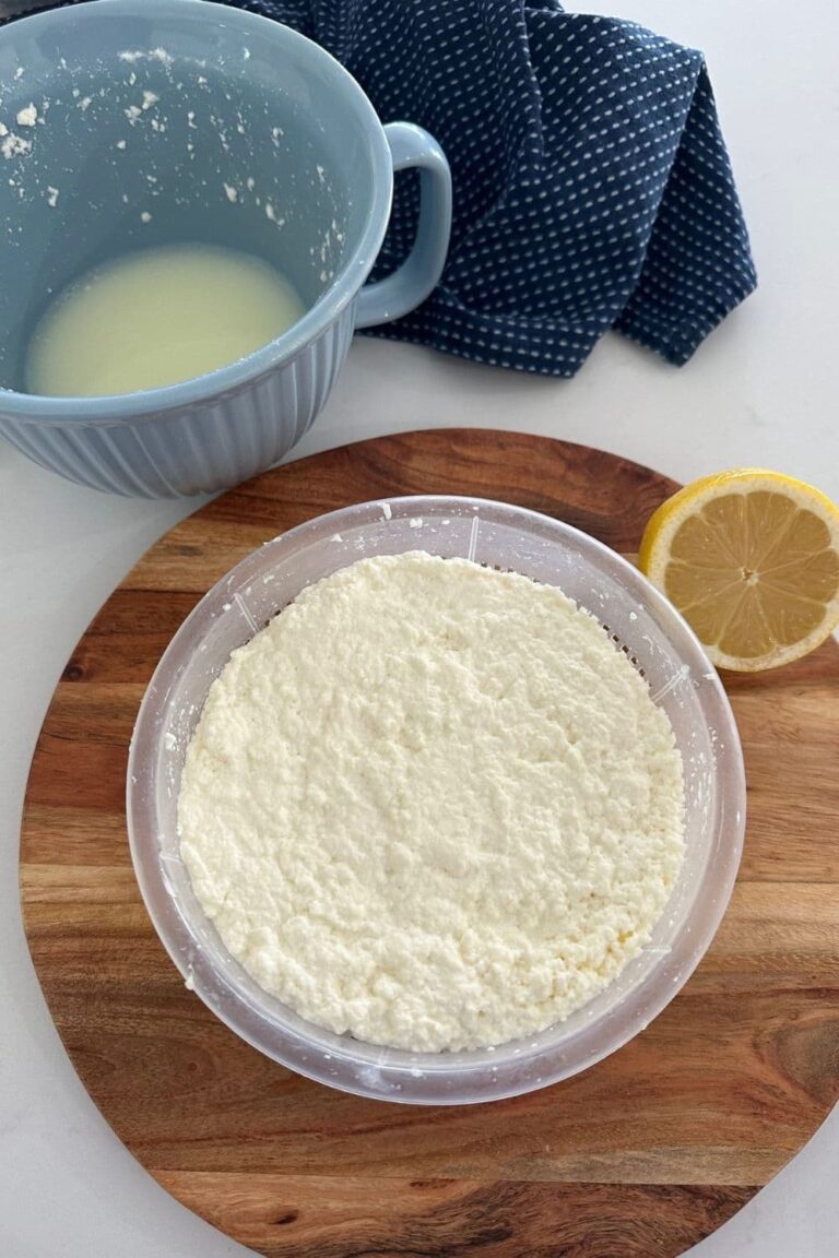 Ricotta cheese that has been strained in a plastic ricotta basket. The basket is sitting on a wooden board and there is a lemon to the right and a blue jug of whey on the left.