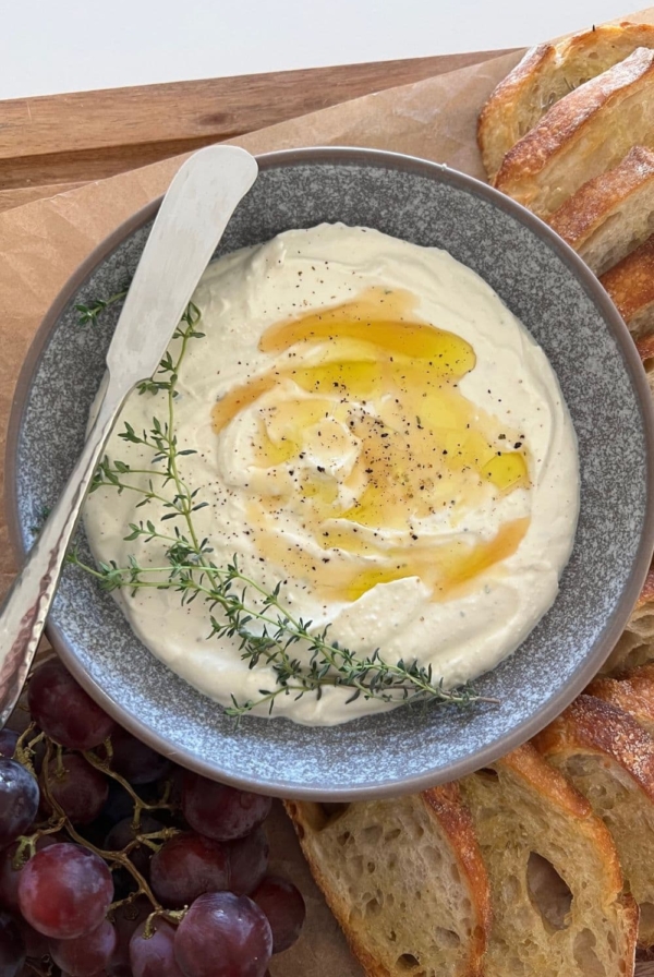 Whipped Ricotta Dip drizzled with honey and olive oil, dressed with fresh thyme leaves and served in a grey stoneware dish. There is a silver pate knife sitting on the edge of the bowl.