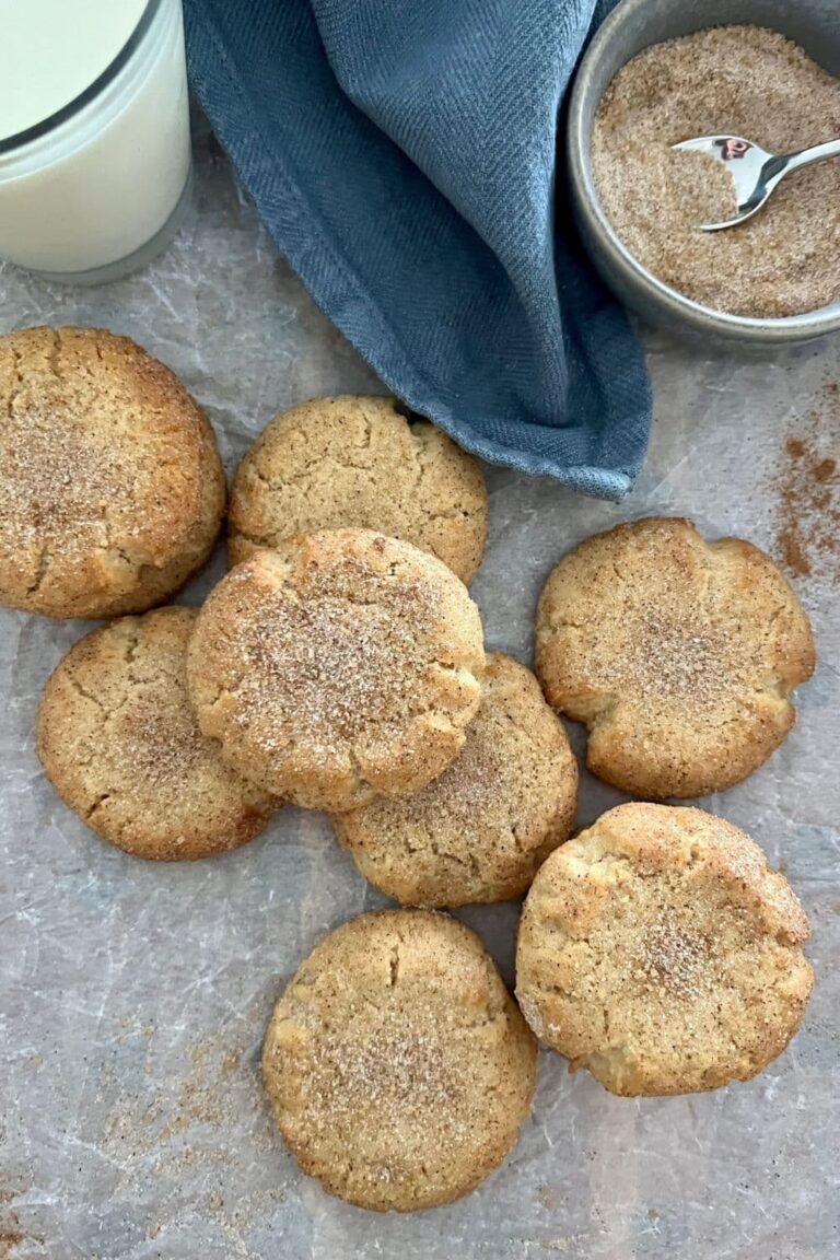 One of the best sourdough cookie recipes - Sourdough snickerdoodles arranged on a piece of parchment paper with a blue dish cloth. There is a bowl of cinnamon sugar in the photo.