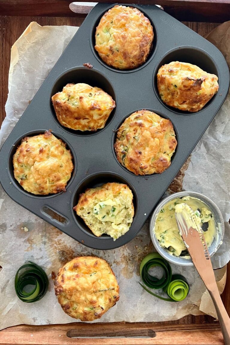 A tray of 6 sourdough zucchini muffins with cheese and chives sitting in a muffin tin. There is a bowl of whipped herb butter sitting next to the sourdough muffins.